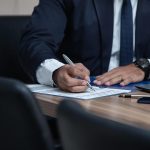 Advogado trabalha ao computador e consulta documentos numa secretária de escritório.