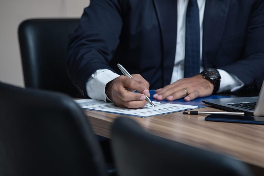 Advogado trabalha ao computador e consulta documentos numa secretária de escritório.